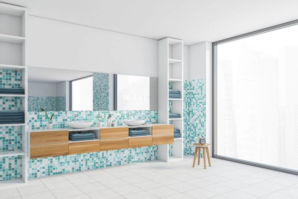 photo of a White and sea-green bathroom with two white sinks and mirror, side view. Wooden drawers and shelves with towels, white tiled floor and window.
