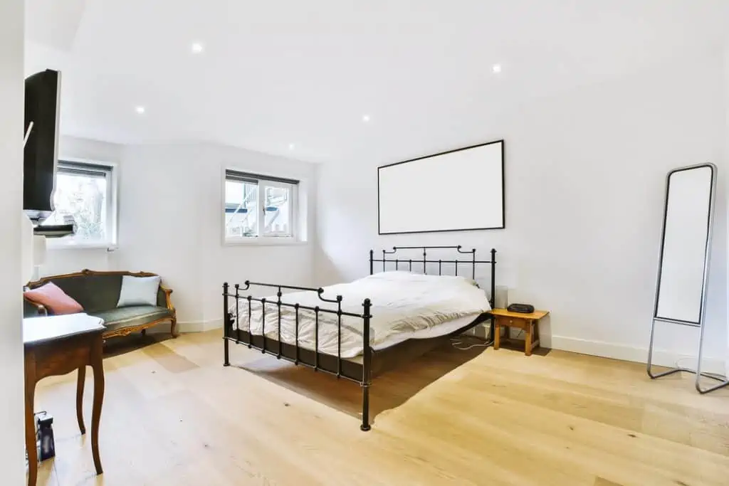 Comfortable bed with black metal frame in room with white wall and wooden floor furnished with vintage sofa and modern mirror